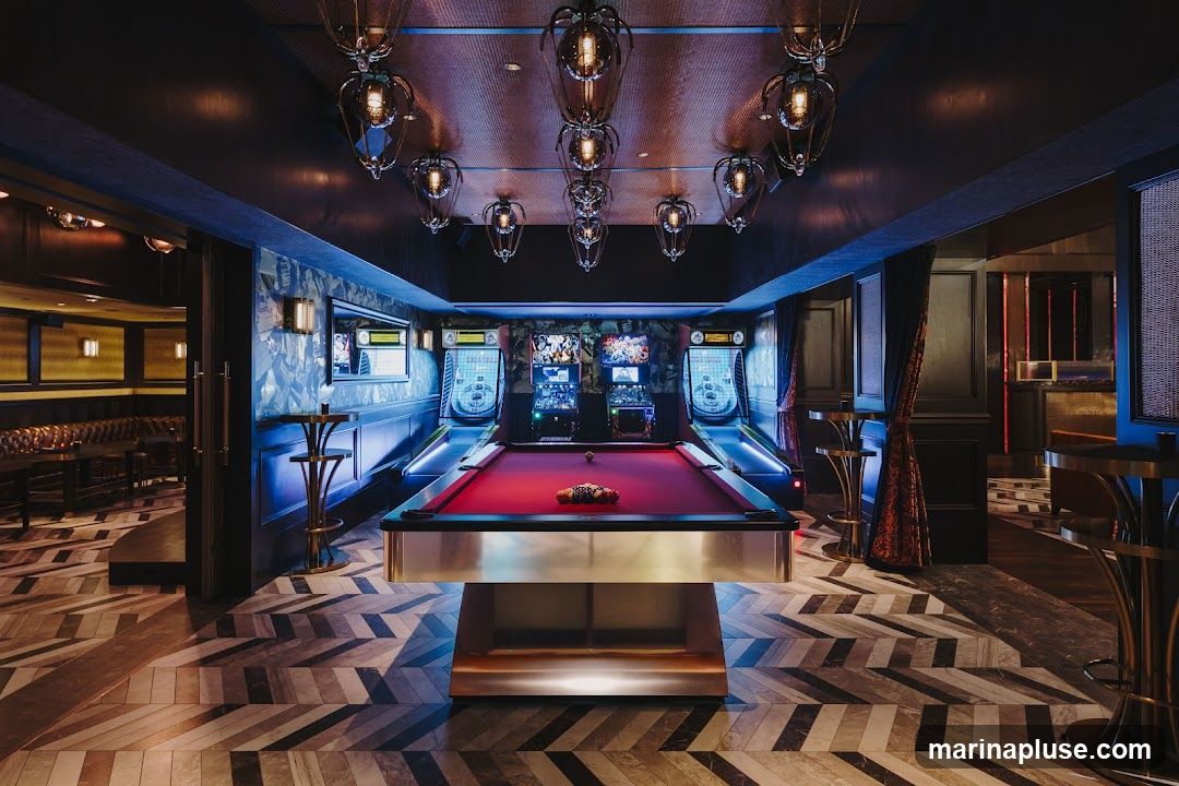 A dimly lit, stylish game room featuring a red felt pool table in the center. Flanking the pool table are two Skee-Ball machines and arcade cabinets. The room has a dark, textured ceiling with ornate pendant lights. The walls are decorated with patterned wallpaper and dark wood paneling. The floor has a distinctive chevron pattern. In the background, a bar area with booth seating is visible.