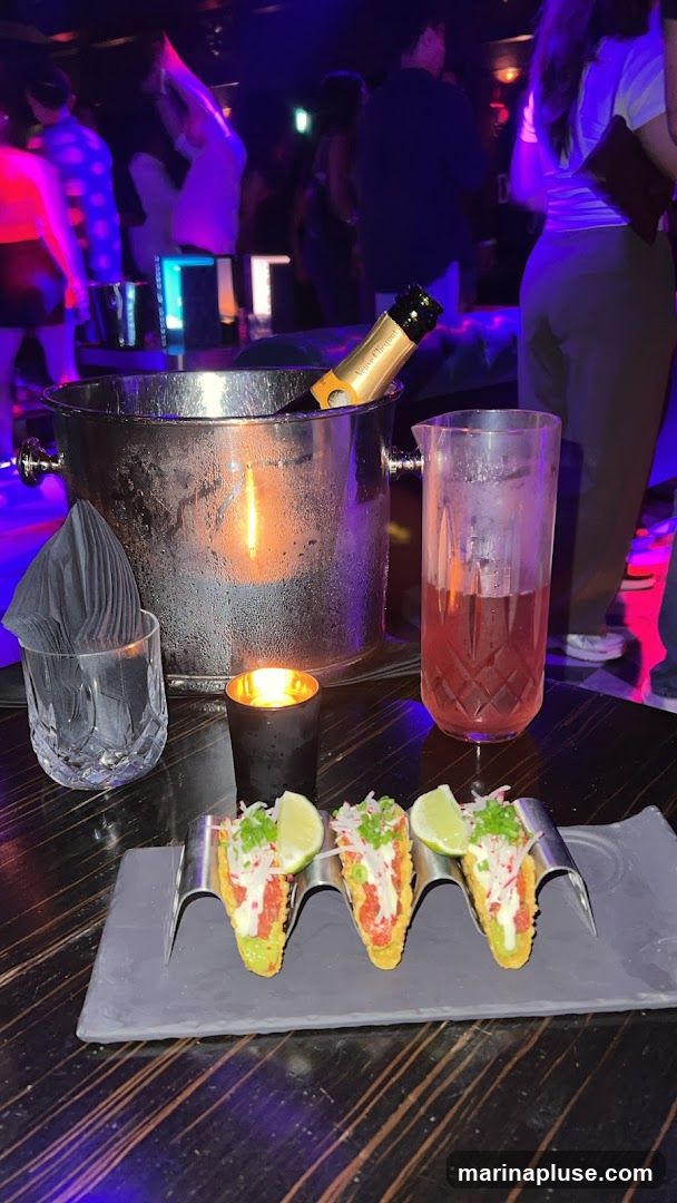 A table at a nightclub with a bottle of champagne in an ice bucket, a cocktail, a candle, and three tacos on a serving tray. People are visible dancing in the background.