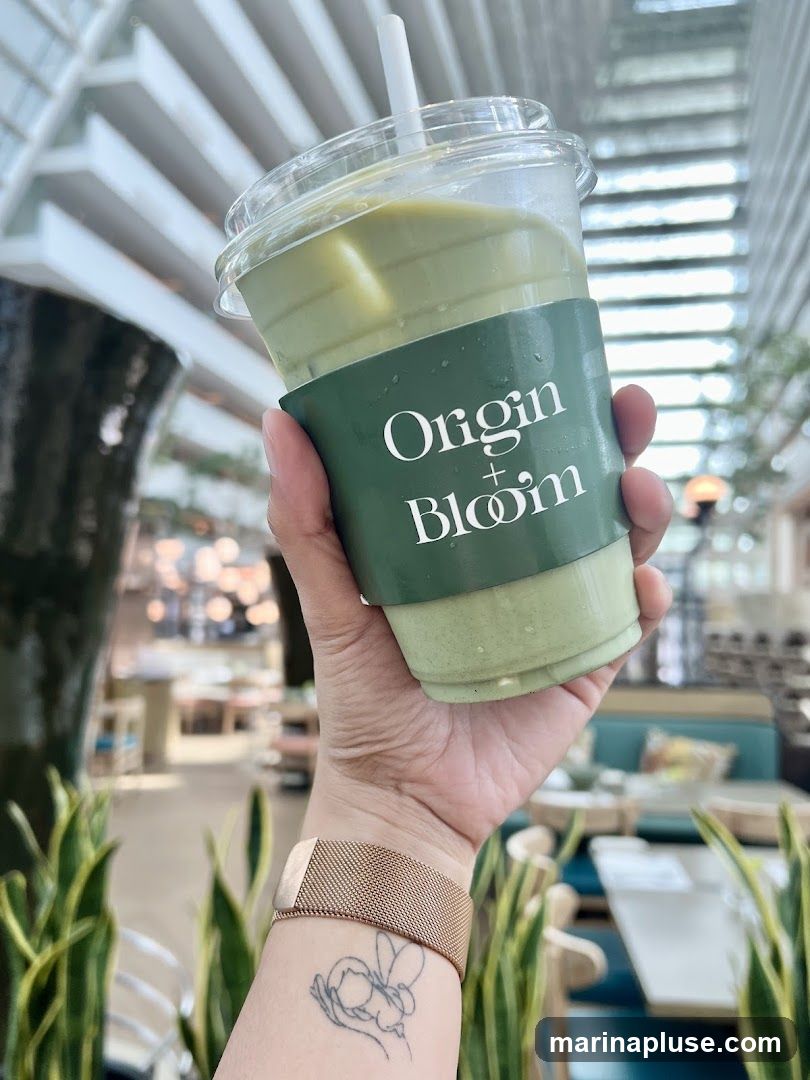 A hand holds a clear plastic cup filled with an iced matcha latte. The cup has a green sleeve with the white text "Origin + Bloom" and a floral logo. The background shows a blurred interior of a cafe with tables, chairs, and plants.