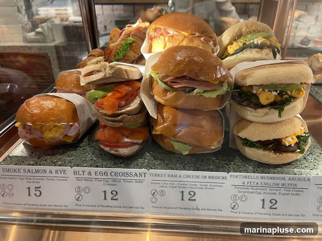 A display of various sandwiches and croissants, including Smoke Salmon & Rye, BLT & Egg Croissant, Turkey Ham & Cheese on Brioche, and Portobello Mushroom, Arugula & Feta English Muffin. Each item has a price listed below its description.