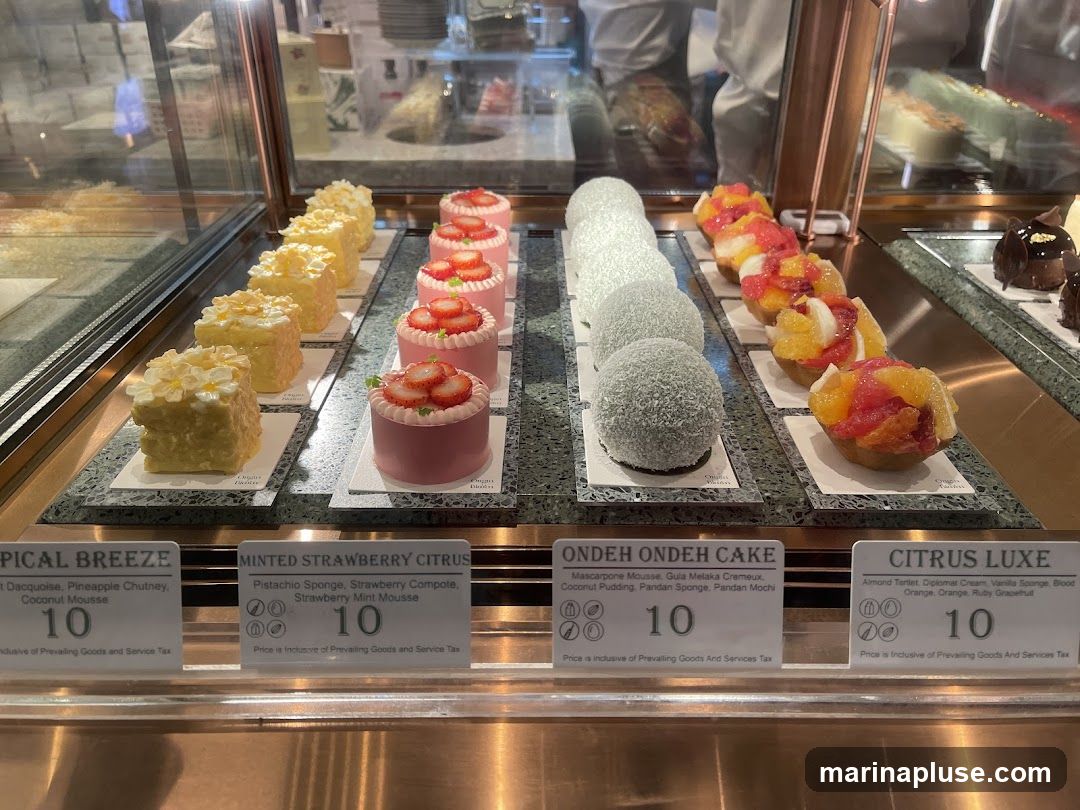 A display case filled with various small cakes and pastries. From left to right, there are yellow cakes topped with white flowers, pink cakes topped with strawberries, round white cakes coated in coconut, and fruit tarts. Each item has a label with its name, ingredients, and price.