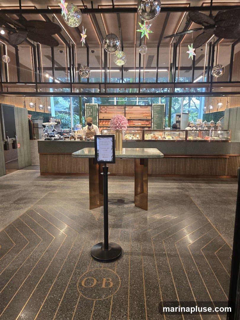 The image shows the interior of a bakery or cafe with a long counter displaying various pastries and breads. A staff member wearing a mask and cap is visible behind the counter. The ceiling is decorated with disco balls and star-shaped ornaments. A sign on a stand in the foreground indicates a queueing area. The floor has a geometric pattern with the letters 'OB' embossed in a circle.