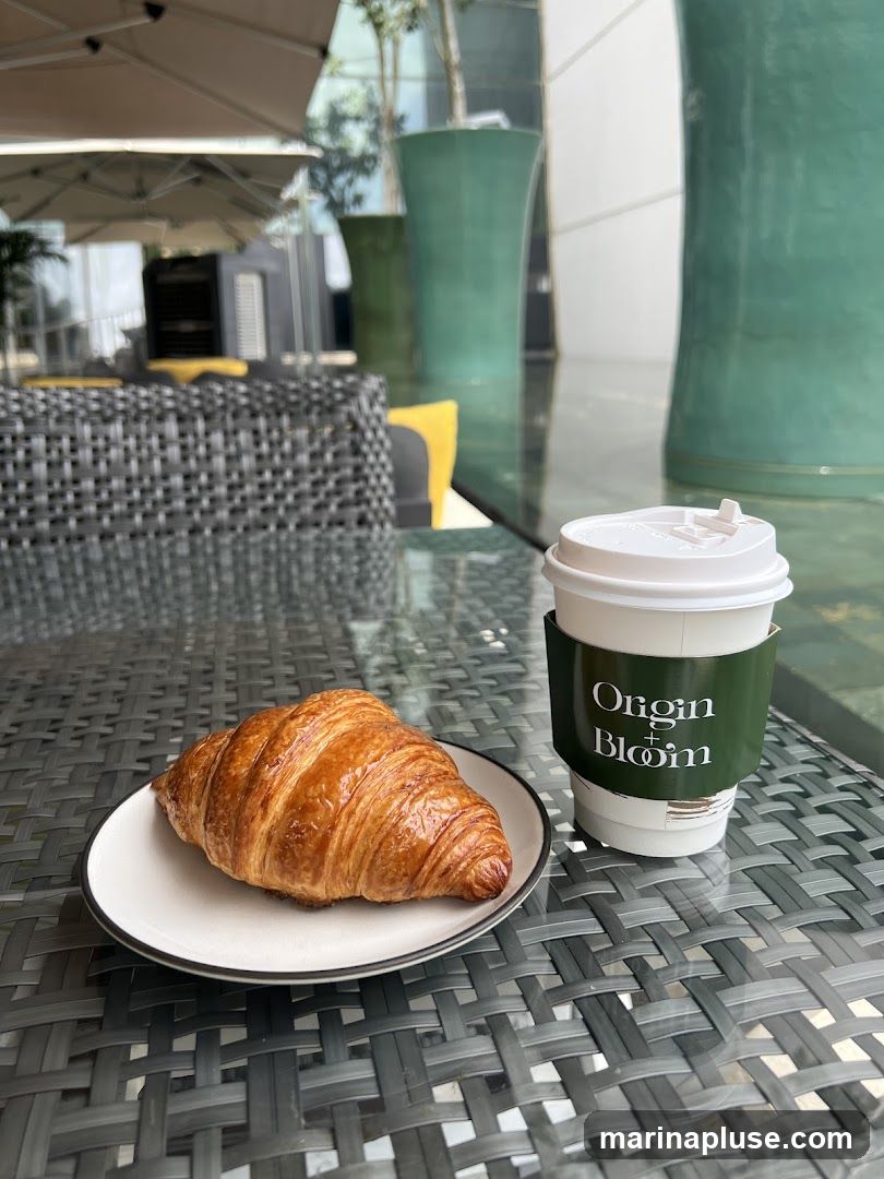 A golden-brown croissant sits on a white plate with a black rim, next to a white coffee cup with a green sleeve. The sleeve has the text "Origin + Bloom" printed in white. The setting appears to be an outdoor cafe with wicker chairs and large green planters in the background.
