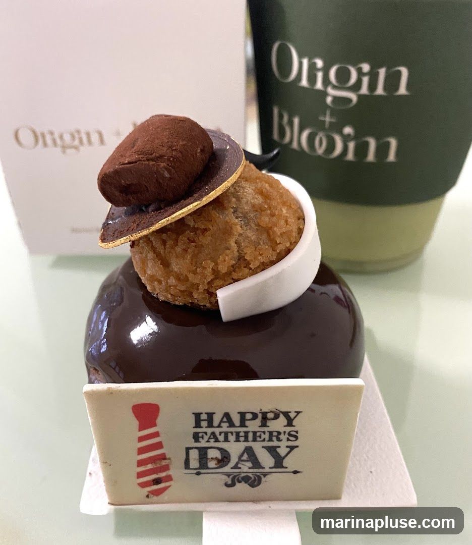 A close-up shot of a chocolate cake decorated to resemble a suit with a tie, presented with a "Happy Father's Day" topper. A green cup with the "Origin + Bloom" logo is visible in the background.