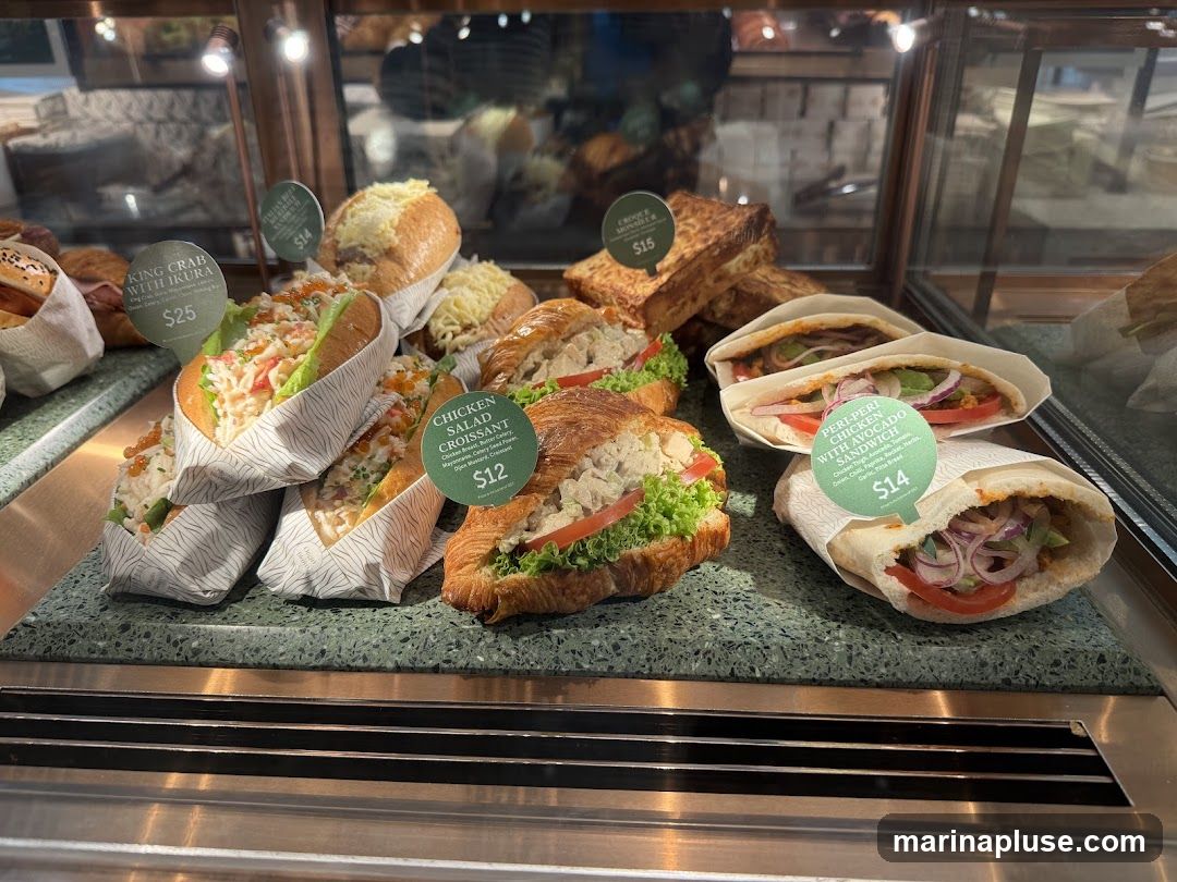 A display case filled with various sandwiches and croissants. Visible items include a King Crab with Ikura roll, Chicken Salad Croissant, and Peri-Peri Chicken with Avocado sandwich. Prices are displayed on circular tags.