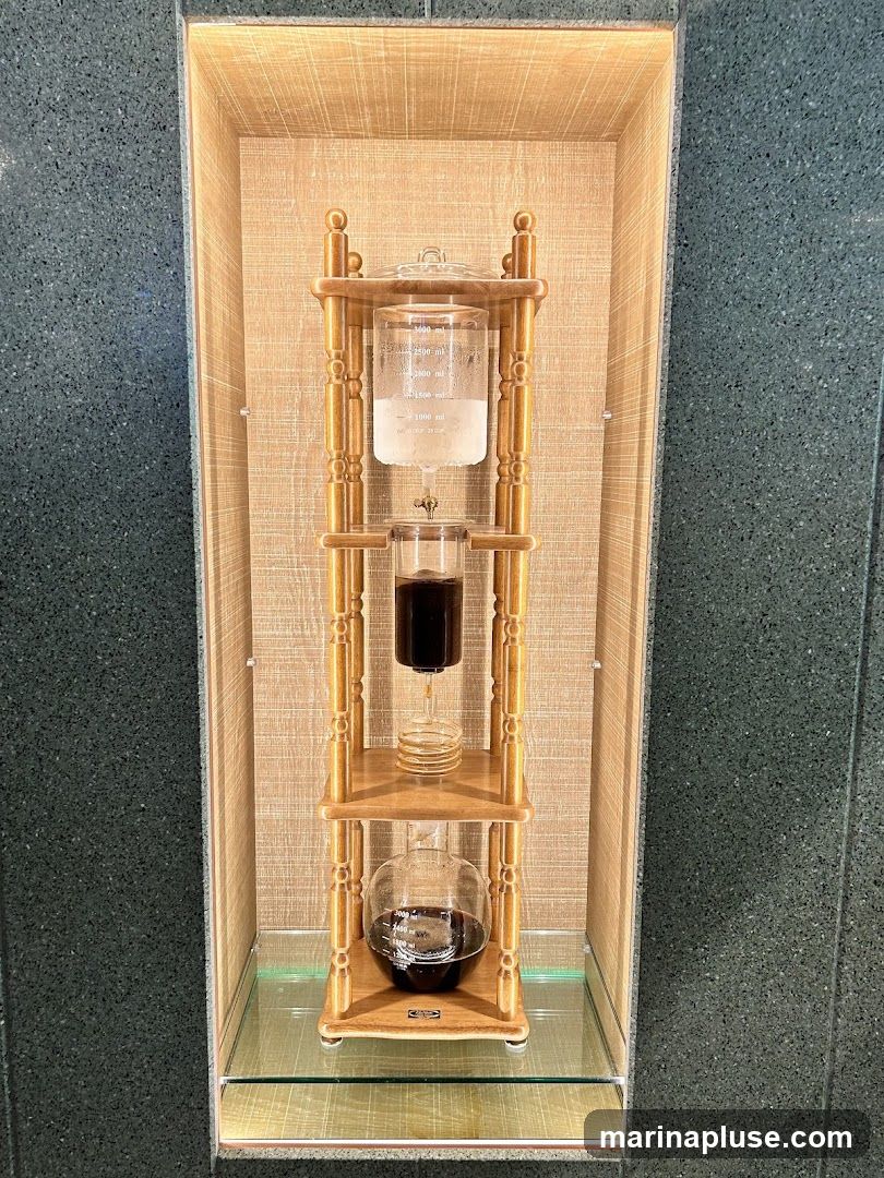 A tall, ornate cold brew coffee maker is displayed in a recessed niche. The maker has three glass chambers, with water in the top chamber, coffee grounds in the middle, and brewed coffee in the bottom. It is made of wood and glass, with a vintage aesthetic. The niche has a textured, light-colored background and is illuminated from above. The surrounding wall is dark and speckled.