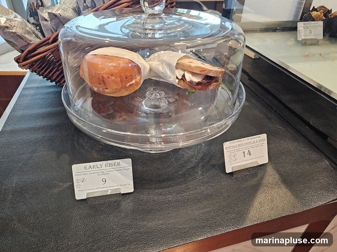 Two sandwiches are displayed under a glass dome on a black surface. To the left is a sandwich labeled "EARLY RISER" with ingredients French Ham, Scrambled Egg, Cheddar Cheese, Pepper, Brioche Bun, priced at 9. To the right is a sandwich labeled "PORTOBELLO MUSHROOM, BUFFALO MOZZARELLA & ROMA" with ingredients Portobello Mushroom, Mozzarella Cheese, Arugula, Balsamic, Caprese Salad, Pesto, Rustic Bread, priced at 14. A basket of bagged items is visible in the background.