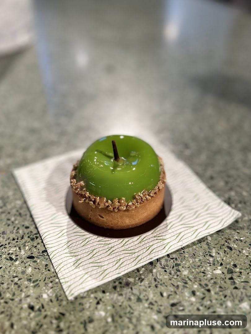 A close-up shot of a green apple-shaped dessert on a white napkin with a green wavy pattern. The dessert has a glossy green glaze and is decorated with gold flakes around the base. The tart sits on a dark brown base and is placed on a speckled green countertop.