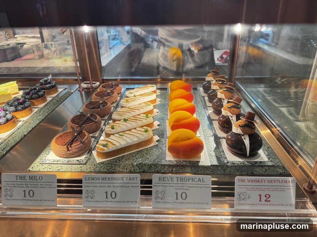 A display case filled with various pastries and desserts. Visible items include tarts topped with blueberries, chocolate mousse cakes, lemon meringue tarts, fruit-shaped mousse desserts, and novelty cakes resembling gentlemen in suits. Labels indicate prices and some ingredients.