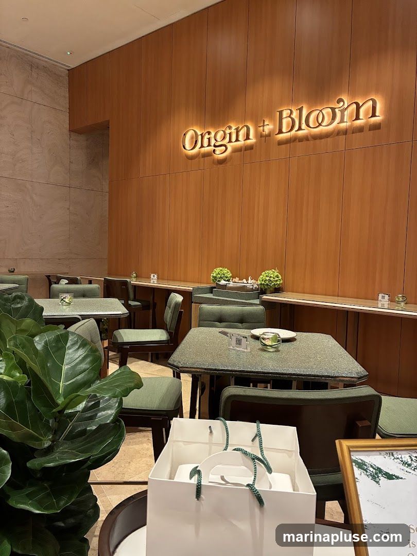 The interior of a cafe or restaurant with a modern and elegant design. The walls are paneled with warm-toned wood, and a prominent illuminated sign reads "Origin + Bloom". The seating area features tables with speckled green tops and chairs upholstered in a matching green fabric. A large potted plant with broad leaves is visible in the foreground, adding a touch of nature to the space. A white gift bag with green handles sits on a table, suggesting a retail or gifting component to the establishment.