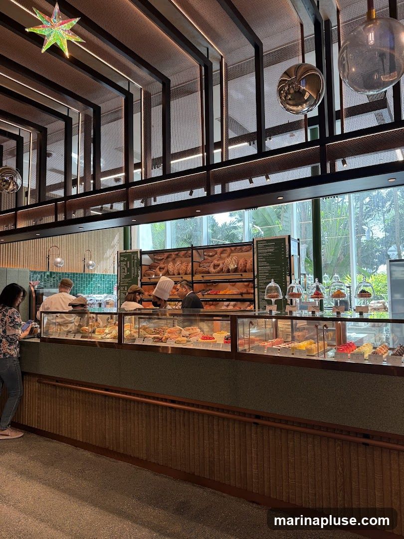 A view of a bakery counter with a variety of pastries and breads on display. Staff members are visible behind the counter, serving customers. The interior features modern lighting and decorative elements.