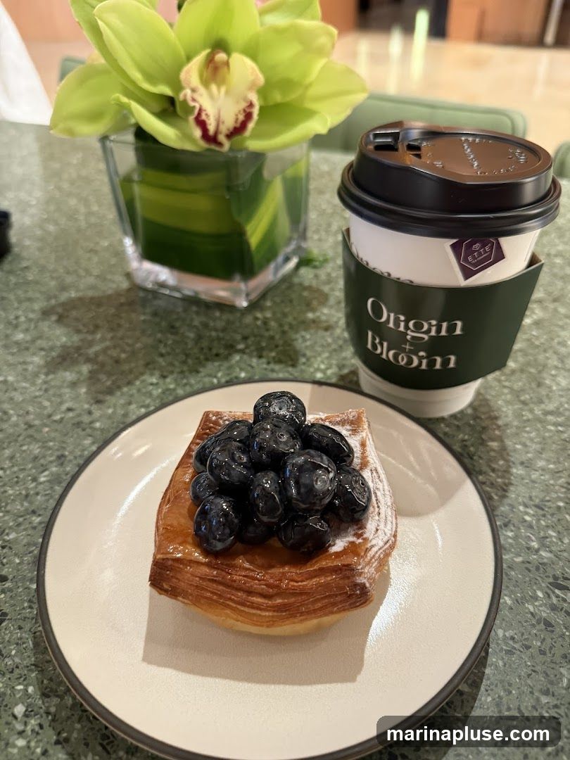 A close-up shot of a blueberry pastry and a cup of coffee on a speckled green table. The pastry is flaky and topped with fresh blueberries and a dusting of powdered sugar. The coffee cup has a green sleeve with the words "Origin Bloom" and a small purple logo. A vase with a vibrant green orchid is visible in the background.