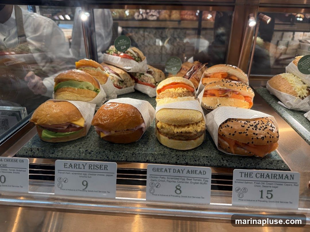 A variety of sandwiches are displayed in a refrigerated case. Some sandwiches are on brioche buns, while others are on bagels. Labels indicate the ingredients and prices of some of the sandwiches.