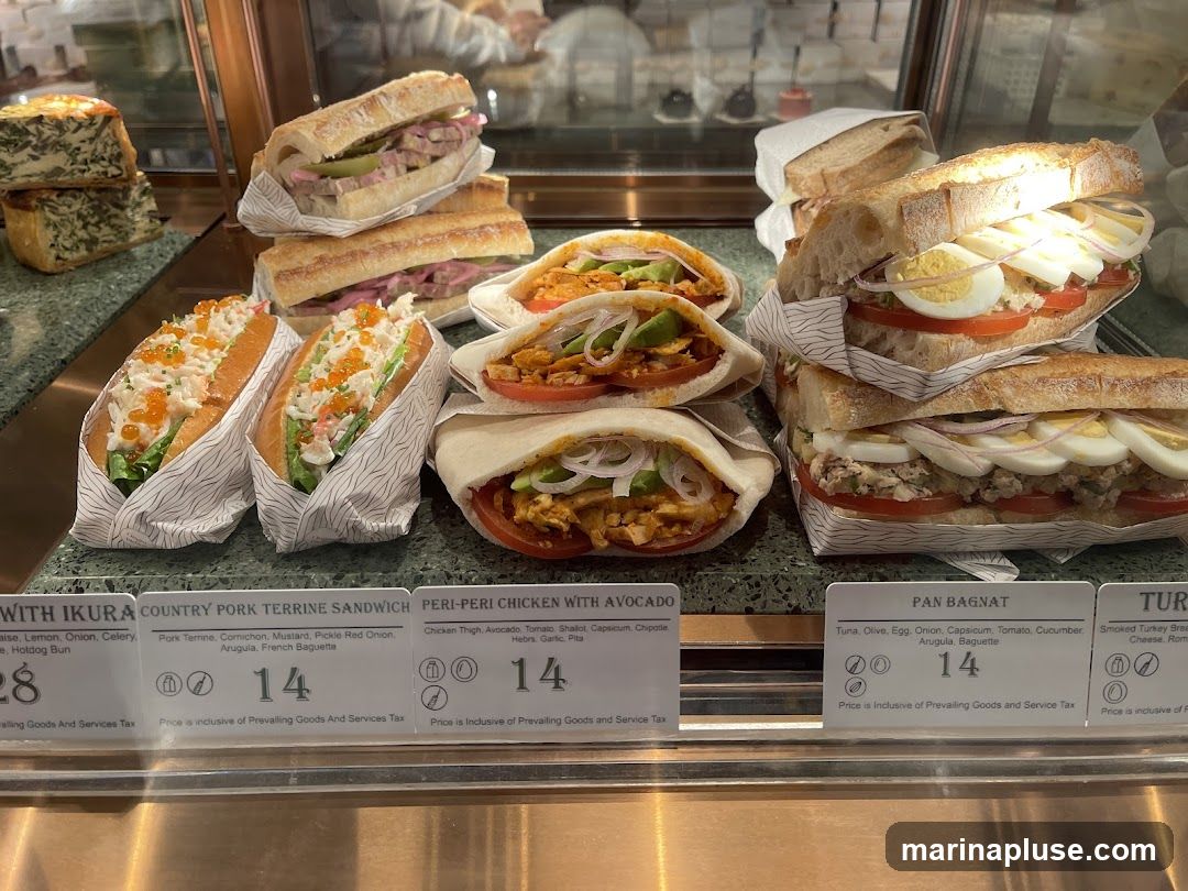 A display of various sandwiches and pitas, including a hotdog bun with ikura, a country pork terrine sandwich, peri-peri chicken with avocado in pita, and pan bagnat. There are also two quiches or savory tarts in the background.