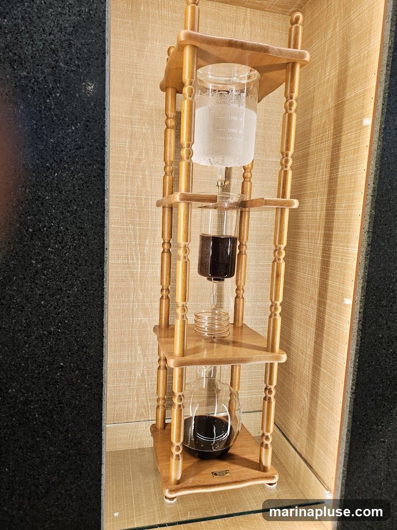 A wooden cold brew coffee maker with three glass chambers. The top chamber contains water, the middle chamber contains coffee grounds, and the bottom chamber contains brewed coffee. The device is displayed on a shelf with a textured background.