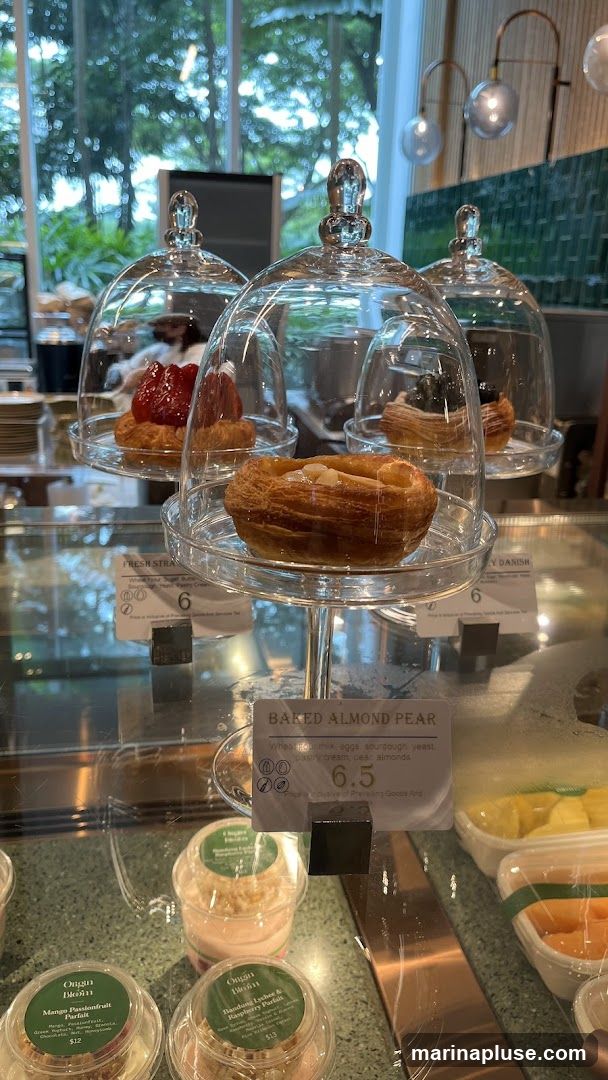 A display case filled with various pastries and parfaits. In the foreground, a baked almond pear pastry is prominently featured under a glass dome. Behind it, a fresh strawberry pastry and a Danish pastry are also visible. Below, several parfaits in clear cups are arranged, including a Mango Passionfruit Parfait and a Bandung Lychee & Raspberry Parfait. The display is set against a backdrop of greenery visible through a large window.
