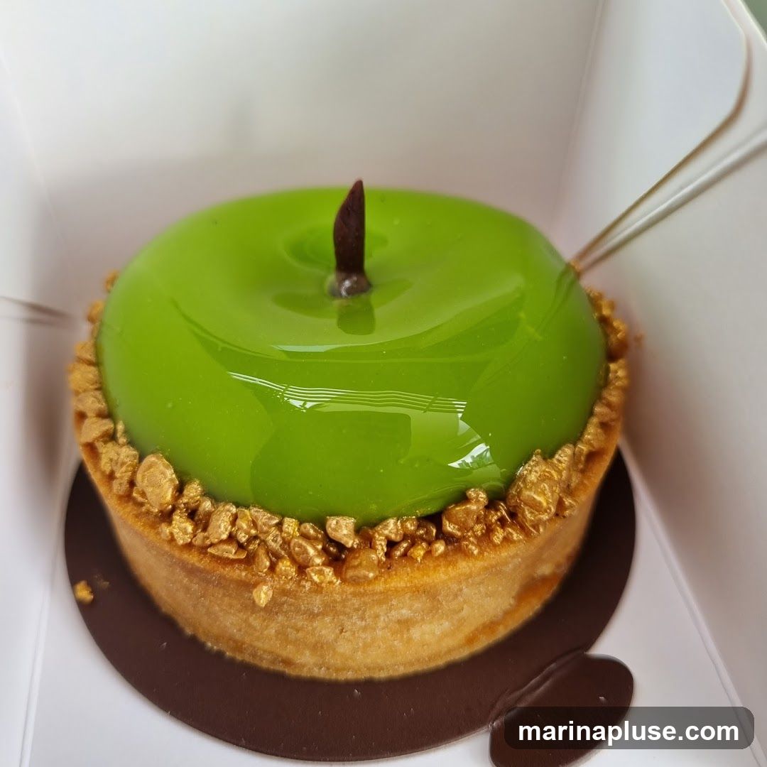 A close-up shot of a green apple-shaped tart in a white box. The tart has a shiny green glaze, a dark brown stem, and a crust decorated with golden nuts.