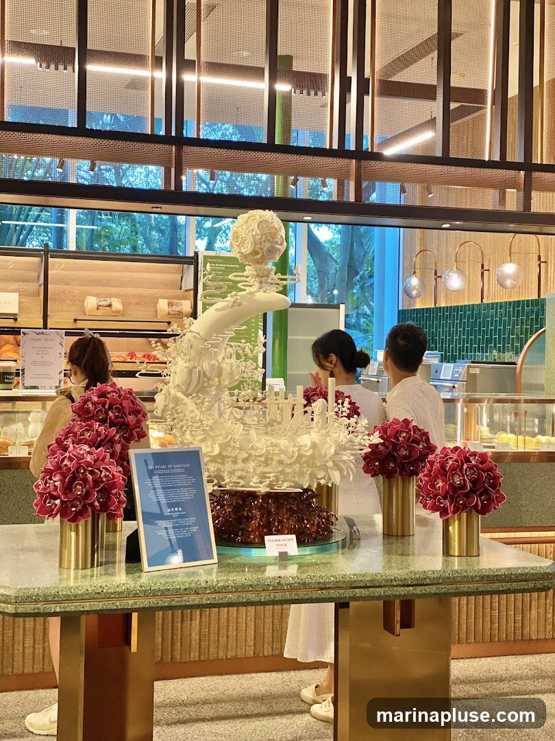 A large, ornate white dessert sculpture is the centerpiece of a display table, surrounded by vibrant red floral arrangements in gold vases. Behind the table, people are visible near a counter with baked goods. The background features a modern interior with decorative lighting and green tiled walls. A sign on the table reads "THE PEARL OF SANYANG" and "PLEASE DO NOT TOUCH.".