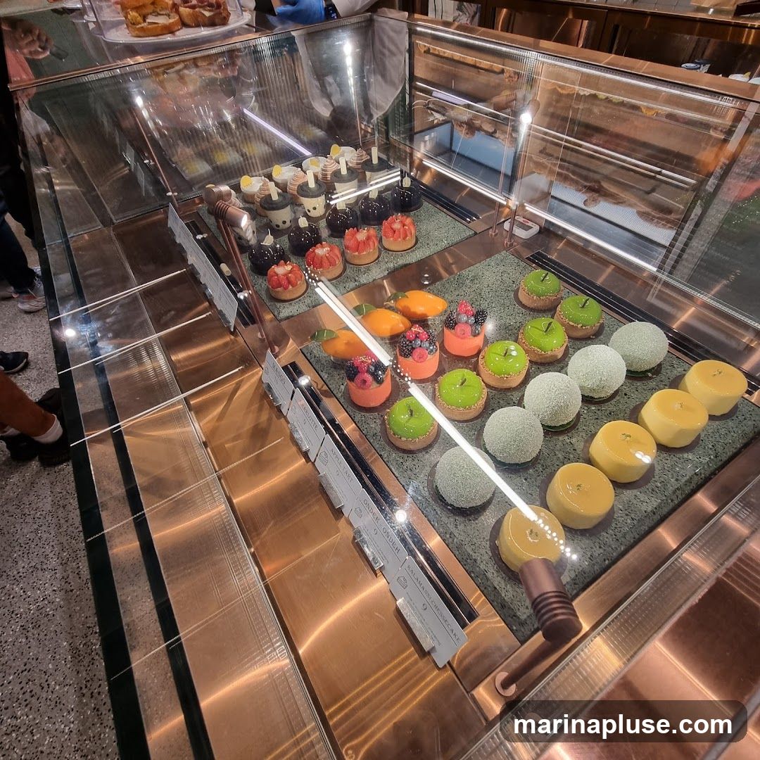 A display case filled with various colorful pastries and desserts. The pastries include fruit tarts, small cakes with green glaze, and round, white-dusted cakes. Some yellow, dome-shaped desserts are also visible.
