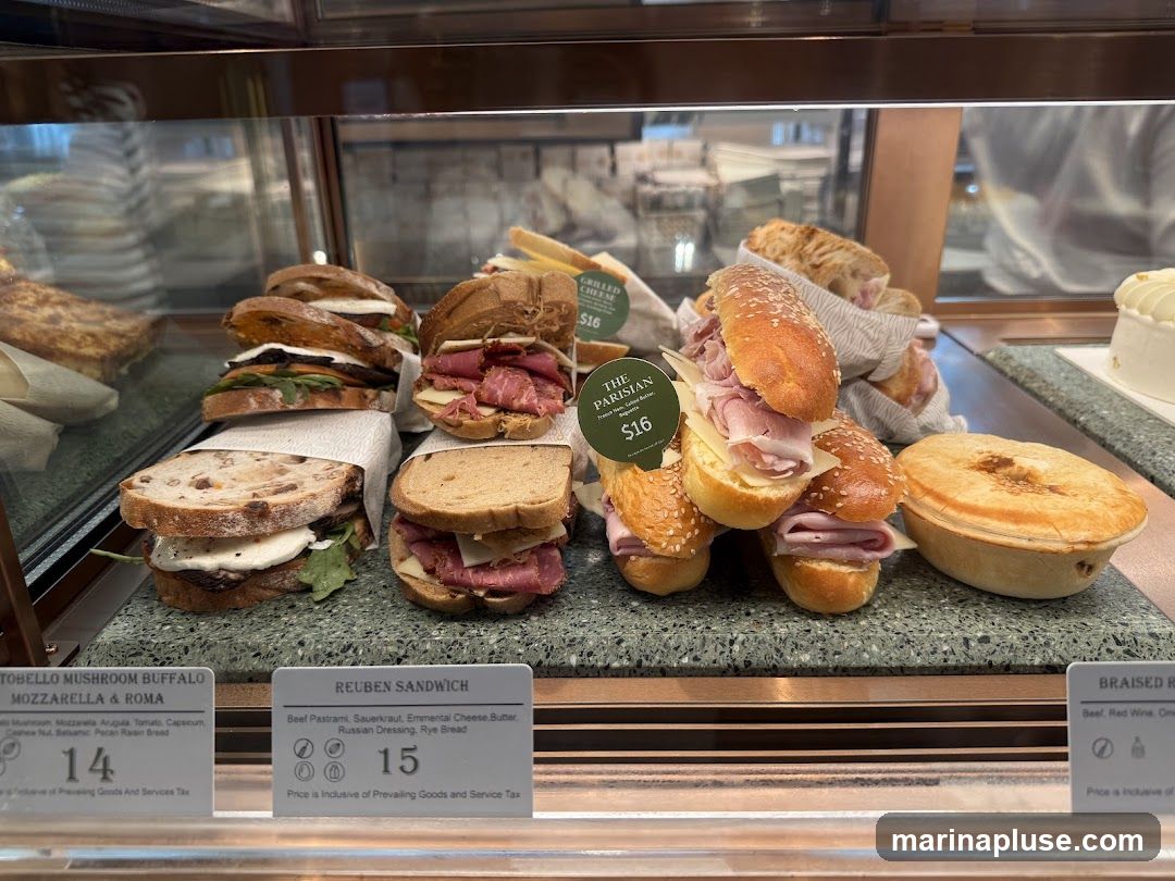 A display case filled with various sandwiches and a pie. In the foreground, there are three labeled items: 'TOBELLO MUSHROOM BUFFALO MOZZARELLA & ROMA' for $14, 'REUBEN SANDWICH' for $15, and a partially visible 'BRAISED R' item. Behind these, several sandwiches are stacked, including ones with pastrami and ham and cheese. A round pie is also visible. A sign for 'THE PARISIAN' with a price of $16 is attached to a stack of ham sandwiches.
