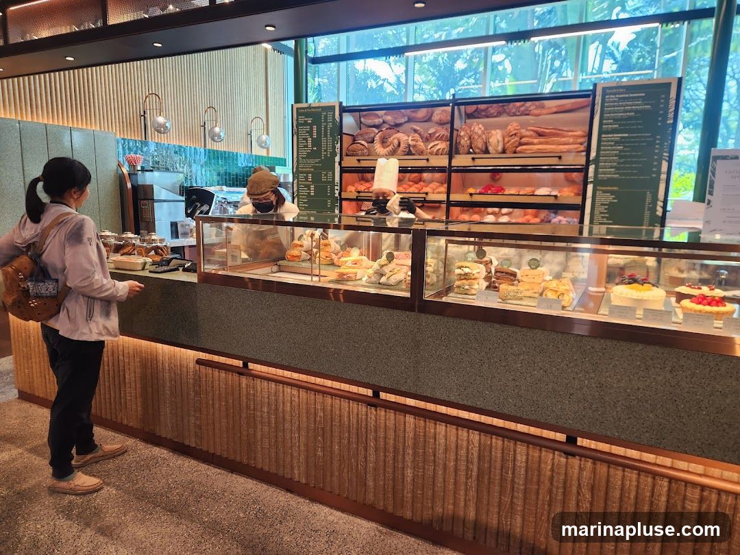 A customer stands at a bakery counter, looking at a display of sandwiches and cakes. Behind the counter, a baker in a chef's hat and mask is visible. Shelves above display various breads and pastries. A menu board is mounted on the wall to the right.