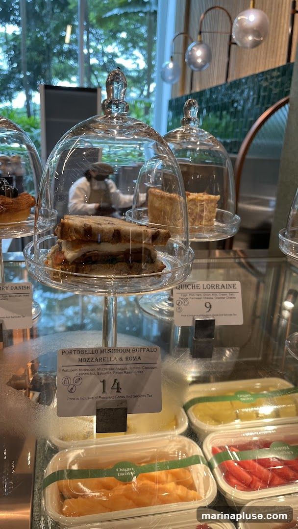 A display case filled with various food items, including sandwiches, quiches, and fruit salads. A sandwich labeled 'Portobello Mushroom Buffalo Mozzarella & Roma' is prominently featured, priced at 14. Another item, 'Quiche Lorraine', is priced at 9. In the background, a person wearing a mask and apron is visible behind the counter.