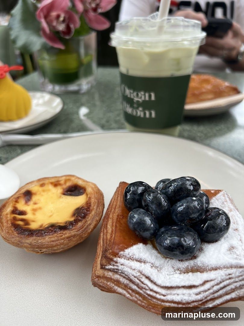 A close-up shot of two pastries and a green drink on a table. The pastry on the left is an egg tart. The pastry on the right is a danish topped with blueberries and powdered sugar. The drink is in a clear plastic cup with a green sleeve that has white text reading "Origin Bloom". In the background, there are pink orchids in a vase and a person holding a phone.