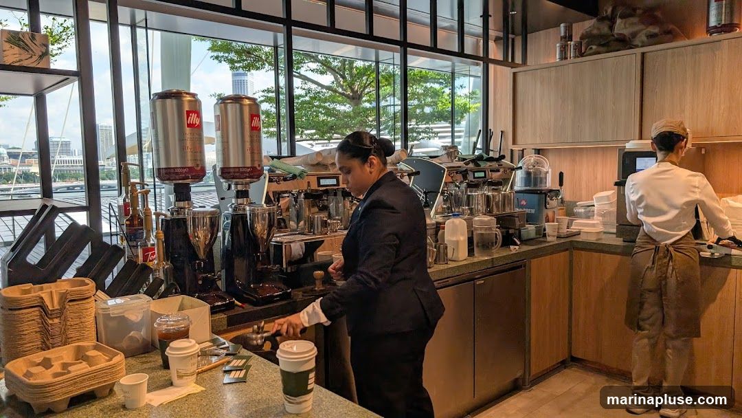 Two baristas are working behind a counter in a modern cafe. The counter is equipped with professional espresso machines and coffee grinders. There are large Illy coffee bean canisters visible. On the left, stacks of cardboard cup holders are neatly arranged. On the right, a barista in an apron is seen from the back, working near a register or payment terminal. The cafe has large windows offering a view of the city skyline and trees.