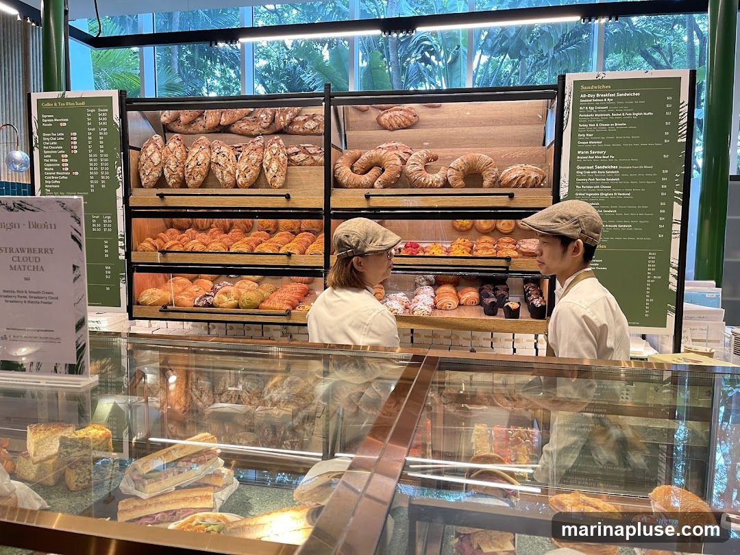 A bakery display case filled with various pastries and sandwiches. Two staff members in uniform and caps are visible behind the counter. Menus are displayed on stands to the left and right of the display.