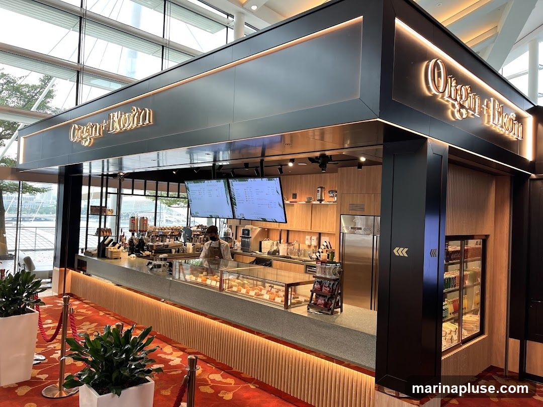 A contemporary coffee kiosk with a sleek black exterior and illuminated signage. The interior features a polished counter with espresso machines, display cases filled with pastries, and a refrigerated section stocked with beverages. Two digital menu boards are mounted above the counter. The kiosk is situated in a bright, open space with large windows and a red carpet with gold accents leading up to it.