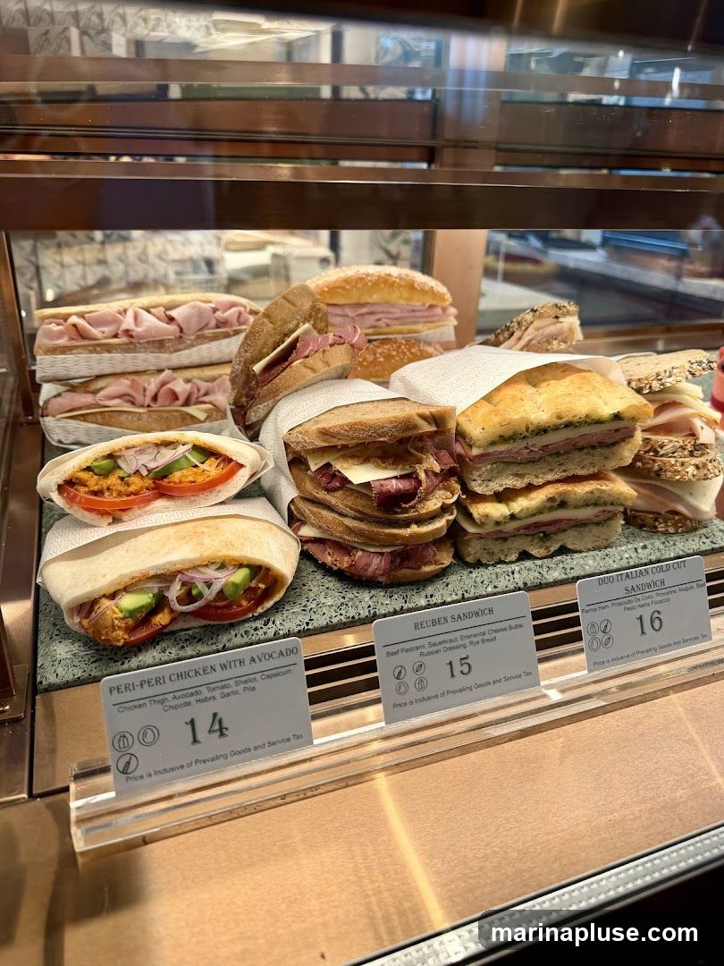 A display case filled with various sandwiches. Visible labels include 'PERI-PERI CHICKEN WITH AVOCADO' for 14, 'REUBEN SANDWICH' for 15, and 'DUO ITALIAN COLD CUT SANDWICH' for 16.