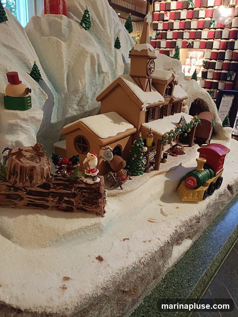 A detailed gingerbread village display with a church, train, and snowy landscape. The village is decorated with miniature Christmas trees, Santa Claus figures, and presents. In the background, a wall is covered with numerous small, individually wrapped cakes or panettone.