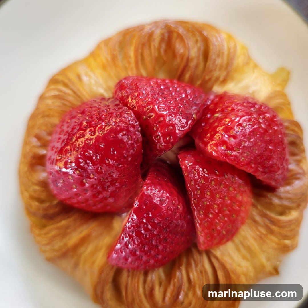 A close-up overhead shot of a golden-brown danish pastry topped with fresh, sliced strawberries. The pastry has a flaky, layered texture, and the strawberries are bright red and glistening.