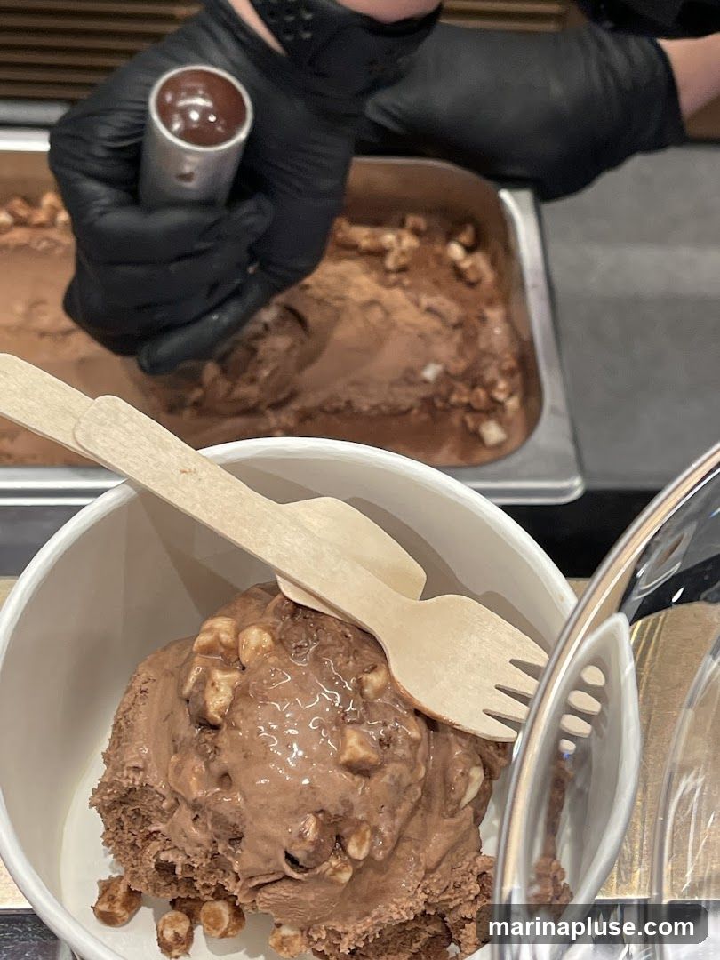 A person wearing black gloves is scooping chocolate ice cream with nuts from a metal container into a white cup. The cup already contains a scoop of ice cream with nuts and two wooden utensils.