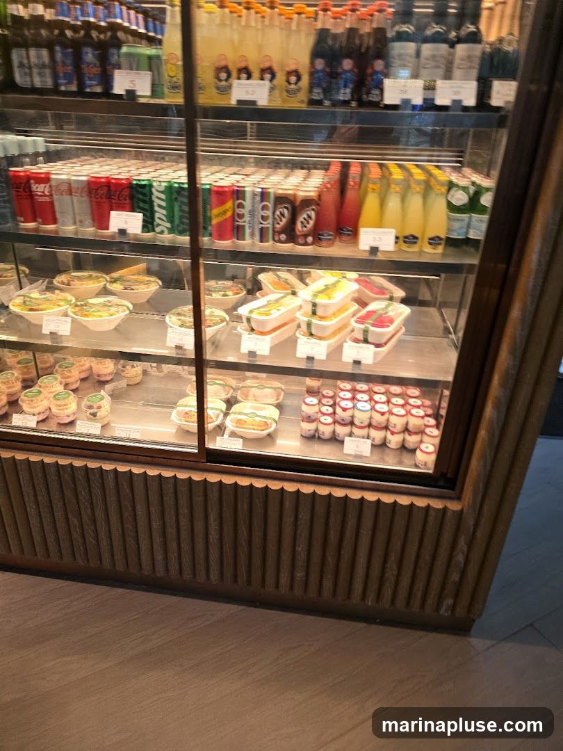 A refrigerated display case filled with various beverages and pre-packaged food items. The top shelf shows bottles of drinks, some with labels like 'Tiger'. The middle shelf is stocked with cans of soda, including Coca-Cola and Sprite, as well as bottles of juice and other drinks. The bottom shelf contains several rows of pre-packaged meals in clear containers, sandwiches, and small yogurt cups. The display case has a wooden base with vertical fluted detailing.