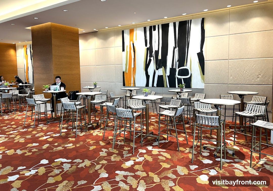 A spacious lounge area with numerous high-top tables and stools, set against a backdrop of textured walls and a large abstract painting. The floor is covered with a vibrant red carpet featuring a floral pattern. Several people are seated at tables, some working on laptops, while others are engaged in conversation. The overall ambiance suggests a contemporary and sophisticated space, possibly a hotel lobby, conference center, or event venue.