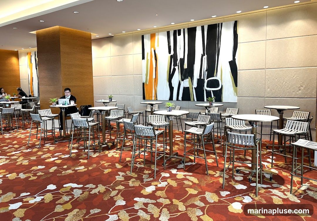 A spacious lounge area with numerous high-top tables and stools, set against a backdrop of textured walls and a large abstract painting. The floor is covered with a vibrant red carpet featuring a floral pattern. Several people are seated at tables, some working on laptops, while others are engaged in conversation. The overall ambiance suggests a contemporary and sophisticated space, possibly a hotel lobby, conference center, or event venue.