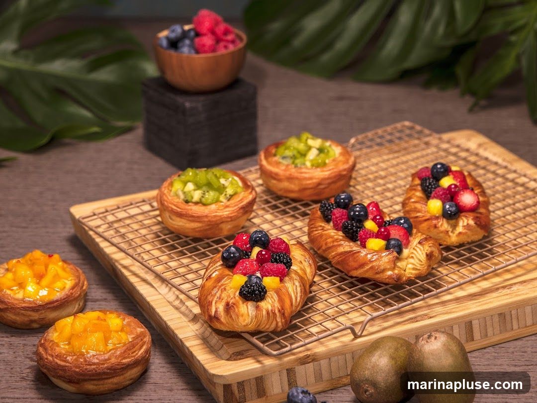 A selection of fruit-filled pastries arranged on a cooling rack and a wooden board. Some pastries are topped with kiwi, while others feature a mix of berries and mango. A small bowl of berries and blueberries is visible in the background, along with whole kiwis.