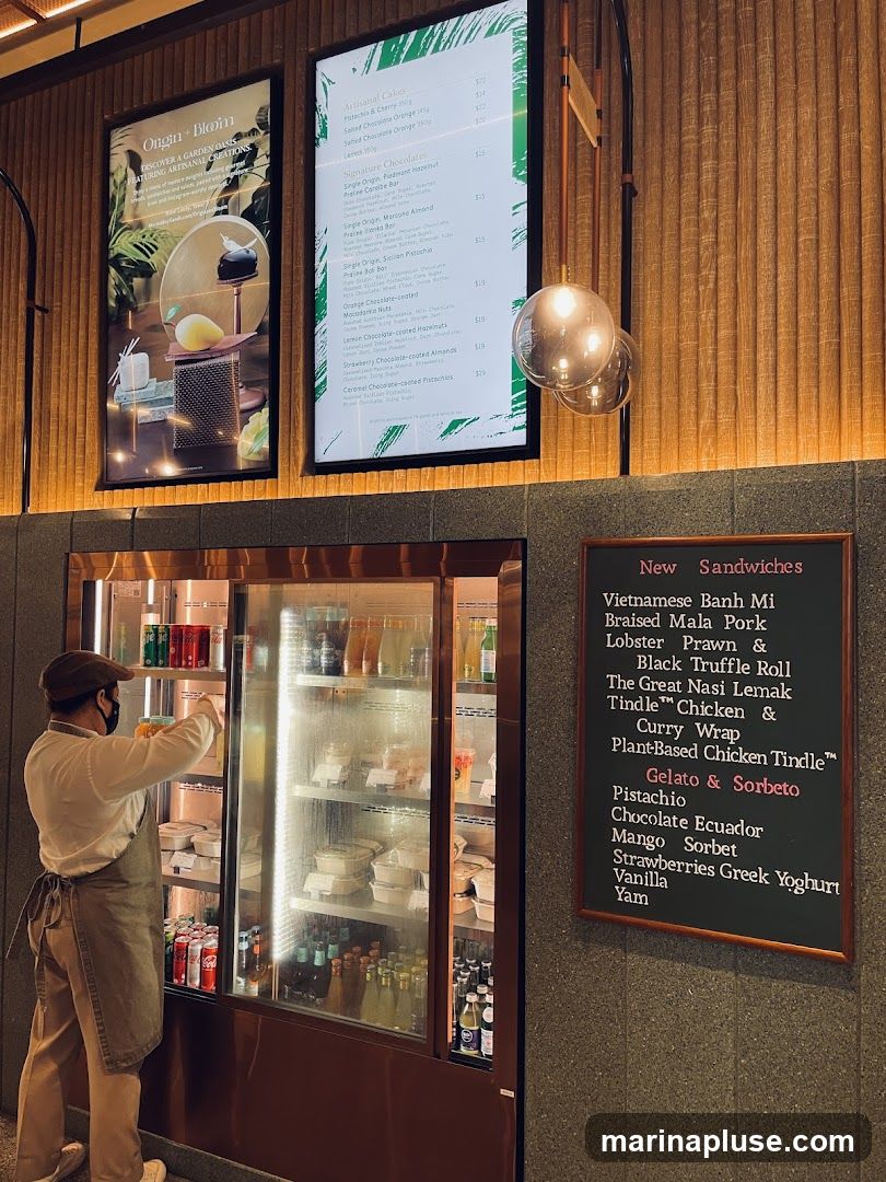 A person in an apron and hat is reaching into a refrigerated display case filled with drinks and pre-packaged food items. Above the display are two illuminated boards: one with an advertisement for "Origin + Bloom" featuring artisanal creations, and another digital menu board listing artisanal cakes and signature chocolates. To the right, a chalkboard menu displays "New Sandwiches" and a selection of gelato and sorbet flavors.