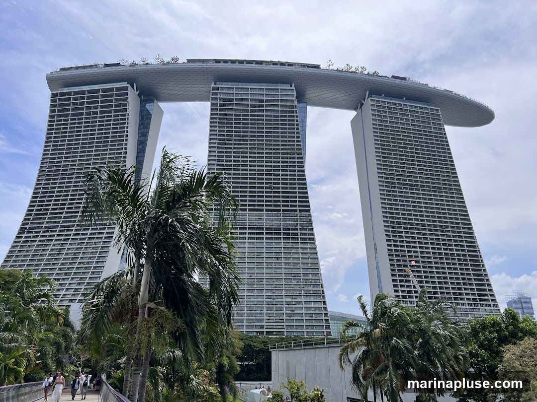 A wide shot of the Marina Bay Sands hotel in Singapore, with its distinctive three towers connected by a large, boat-shaped rooftop structure. Palm trees and lush greenery are in the foreground, and a walkway with people can be seen on the left. The sky is partly cloudy.