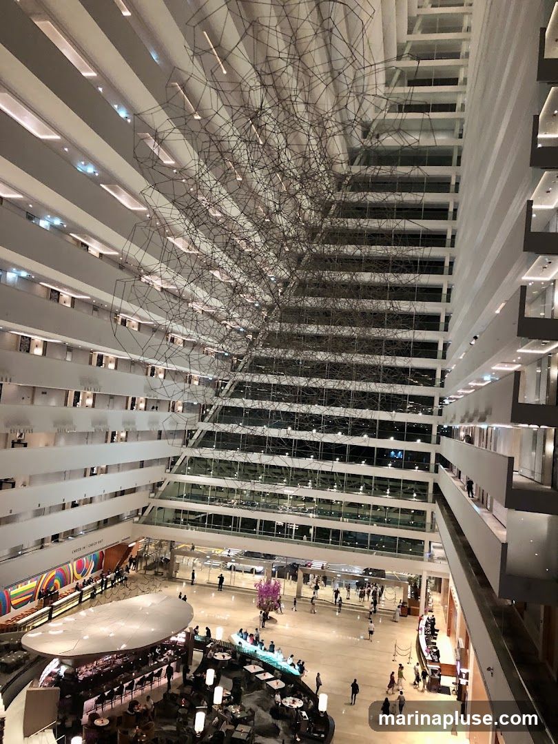 A wide-angle view from above of a large, multi-story hotel atrium. A complex, wireframe sculpture hangs from the ceiling. The atrium features multiple levels of balconies with rooms, a ground floor with a bar, seating areas, and a reception desk. People are visible walking around the ground floor and seated at the bar and tables.