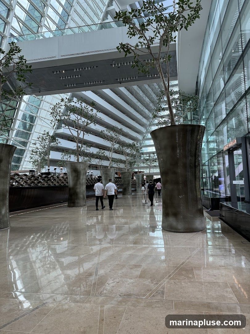 A wide, polished marble floor reflects the light in a spacious, modern atrium. Large, dark planters with tall, slender trees are strategically placed throughout the area. The architecture features a striking glass facade on the right and a multi-tiered, angular structure on the left, creating a sense of depth and grandeur. Several people are walking through the atrium, some in white shirts, suggesting a hotel or corporate environment. The overall impression is one of contemporary elegance and open space.