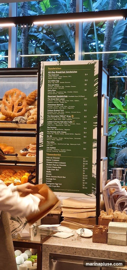 A menu board displays a selection of sandwiches and viennoiseries with prices. The background shows a display of baked goods and lush greenery through a window.