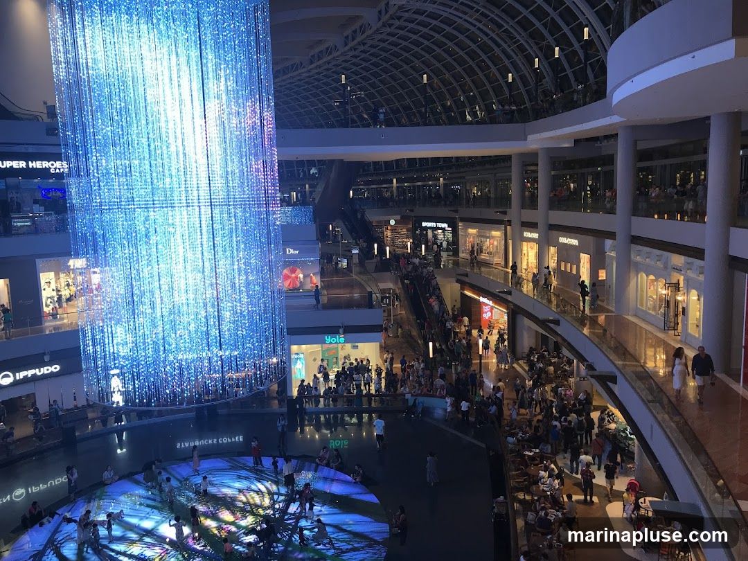 The image shows the interior of a multi-level shopping mall. A large, illuminated art installation made of blue lights hangs from the ceiling, dominating the left side of the frame. Below it, a circular area with projected light patterns on the floor is visible, with people gathered around. Various shops with visible signage like 'IPPUDO', 'SUPER HEROES CAFE', 'DIOR', 'YOLO', and 'SPORTS TOWN' are seen on different levels. Escalators and walkways are filled with shoppers, indicating a bustling atmosphere.
