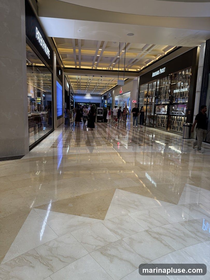 A wide, well-lit interior of a luxury shopping mall with polished marble floors reflecting the overhead lighting and storefronts. Several high-end watch retailers, including Patek Philippe and Audemars Piguet, are visible. Shoppers are seen walking down the corridor.