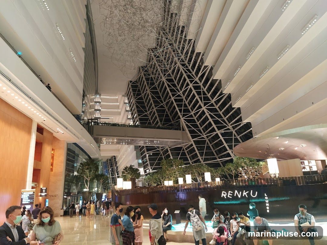 The image shows the interior of a large, modern building with a high ceiling and multiple levels. A distinctive geometric structure is visible in the background. In the foreground, people are gathered, some standing and some sitting. A sign that reads "RENKU LOUNGE" is visible on a dark, curved structure. The overall ambiance is one of a public space, possibly a hotel lobby or a shopping mall, with contemporary architecture and lighting.