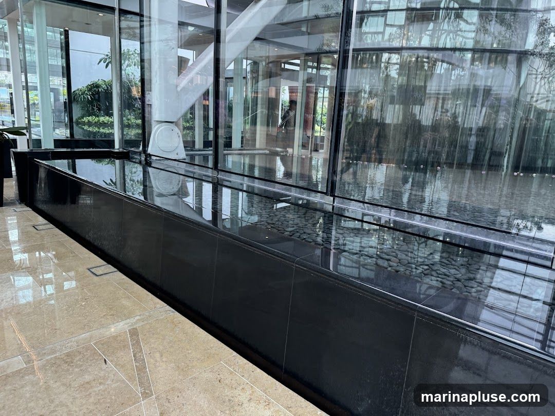 A contemporary interior space featuring a long, black-tiled water feature filled with pebbles and water. The water reflects the surrounding environment, including large glass windows, architectural elements like a white beam and staircase, and greenery visible through the glass. The floor is tiled with large, light-colored stone tiles.