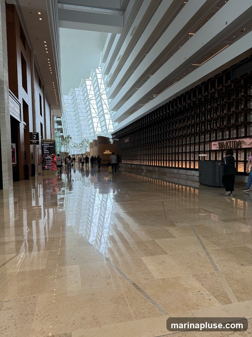 A wide shot of a modern, multi-level interior atrium with a polished marble floor reflecting the architecture. The left side features wooden paneling and signage, while the right has a dark, textured wall with illuminated panels. A striking geometric glass structure dominates the background, with people walking through the space.