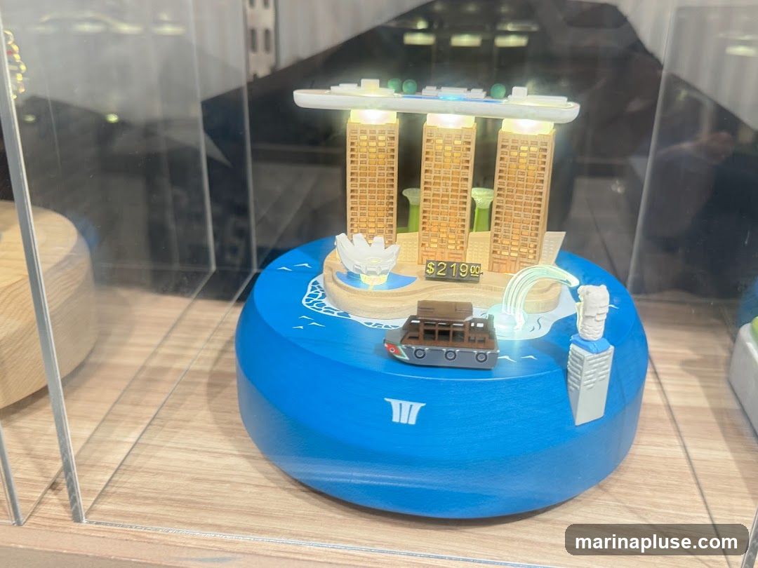 A miniature replica of the Marina Bay Sands hotel and Merlion statue, displayed on a blue base with a toy boat. The replica is illuminated and has a price tag of $219.00. It is housed in a glass display case.