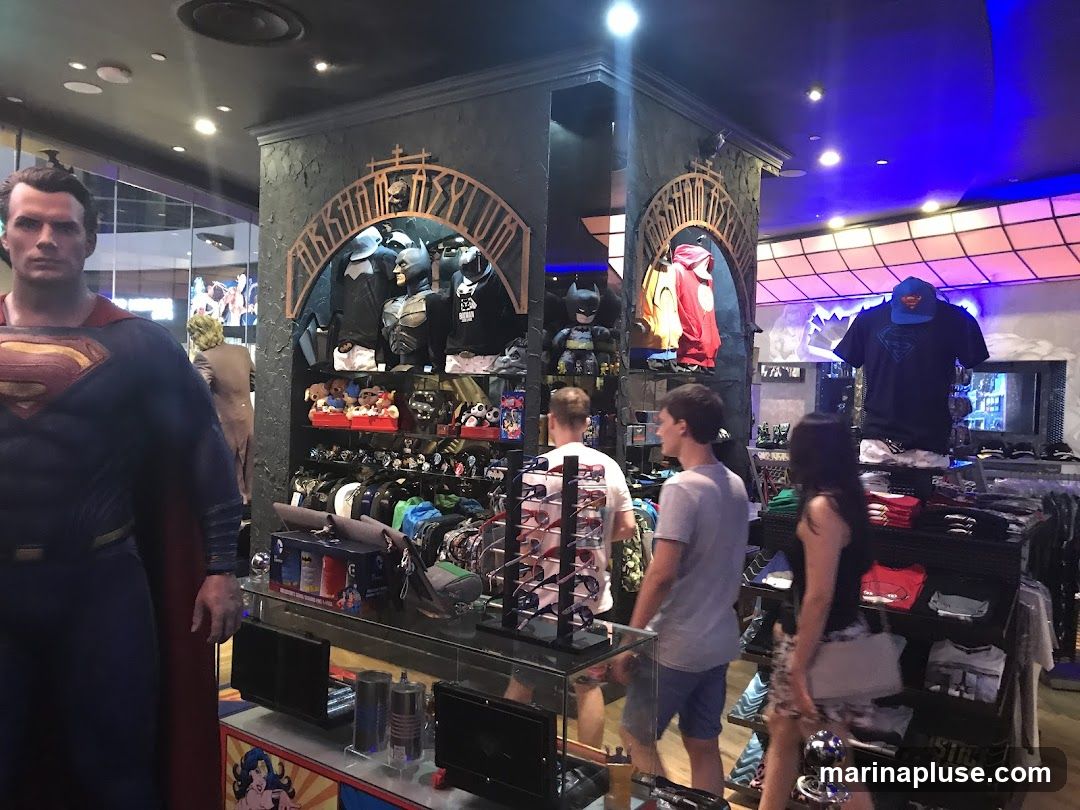 A store interior filled with merchandise related to comic book characters, primarily Batman and Superman. A large Superman statue stands prominently in the foreground. Shelves display various items including costumes, t-shirts, plush toys, and accessories. Several customers are browsing the merchandise. The store features themed decor, including an archway with the word "Arkham Asylum" visible.